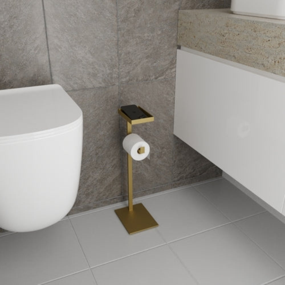 Fayanslı duvarlara ve şık yüzeylere sahip modern bir banyo ortamında sergilenen, aksesuarlar için küçük bir rafa sahip altın rengi, yere monte bir tuvalet kağıdı tutucusu.