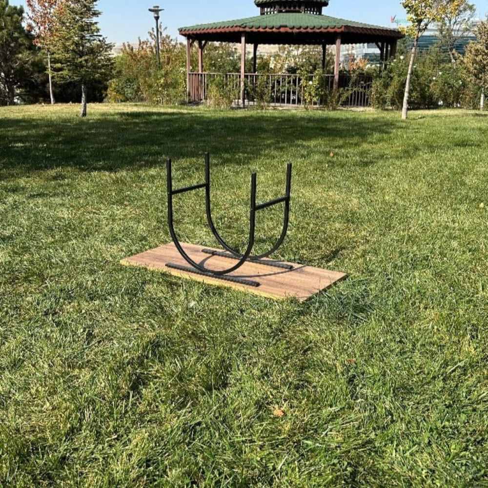 Arka planda yeşil çimler ve bir kameriyenin bulunduğu bir açık hava parkında, ahşap bir kalas üzerine yerleştirilmiş, içinde odun bulunmayan siyah oval şeklinde metal bir odunluk.