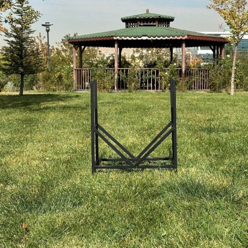 Arka planda manzaralı bir kameriye görüntüsü olan, yeşil çimlerin üzerinde dış mekanda yer alan, V şeklinde tasarıma sahip, boş siyah metal geometrik bir odunluk.