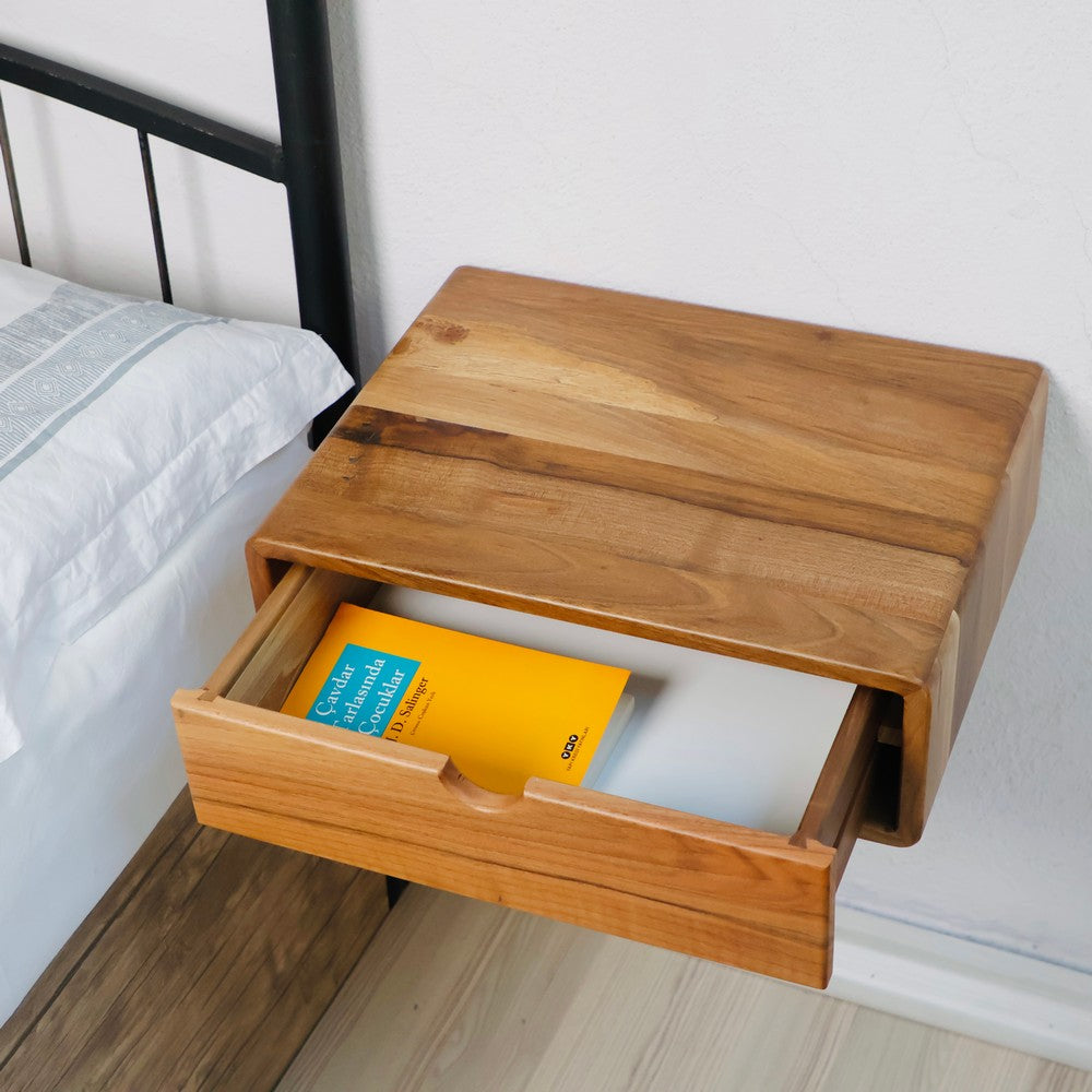 Duvara monte ahşap komodinin çekmecesi açık, içinde bir kitap görünüyor. Doğal ahşap dokusu ve kompakt tasarımı, minimalist yatak odası düzenlemeleri için idealdir.