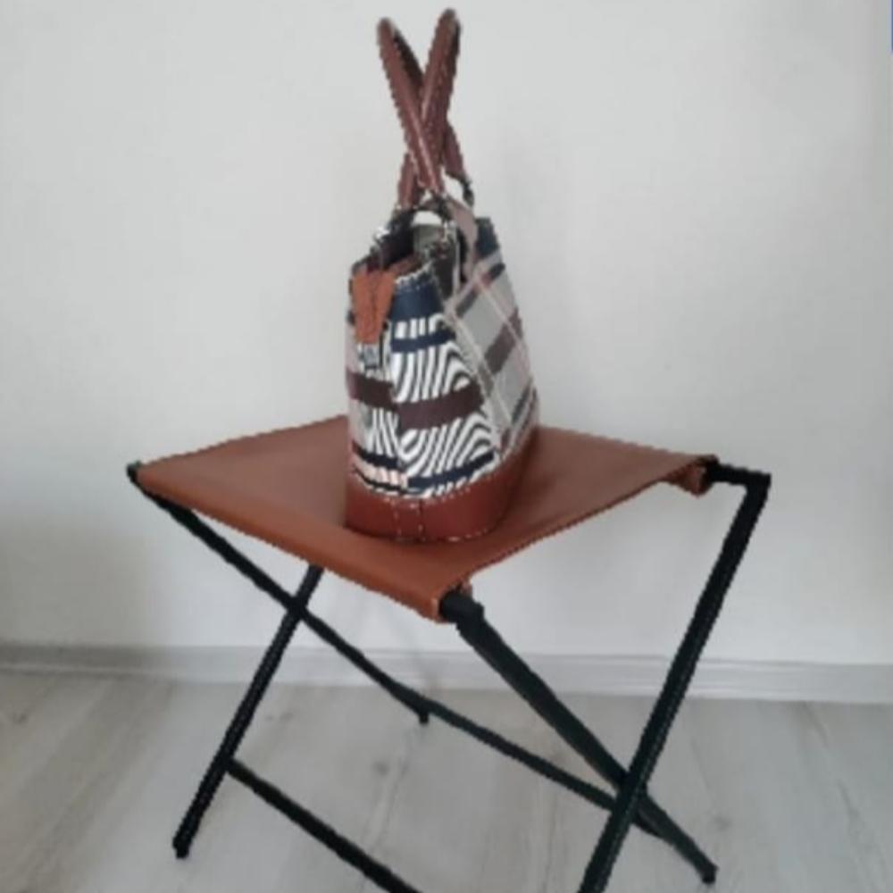 Açık renkli ahşap bir zeminde duran, hakiki deri yüzeyli katlanabilir metal bir bavul rafının üzerine yerleştirilmiş şık ekose bir el çantası.