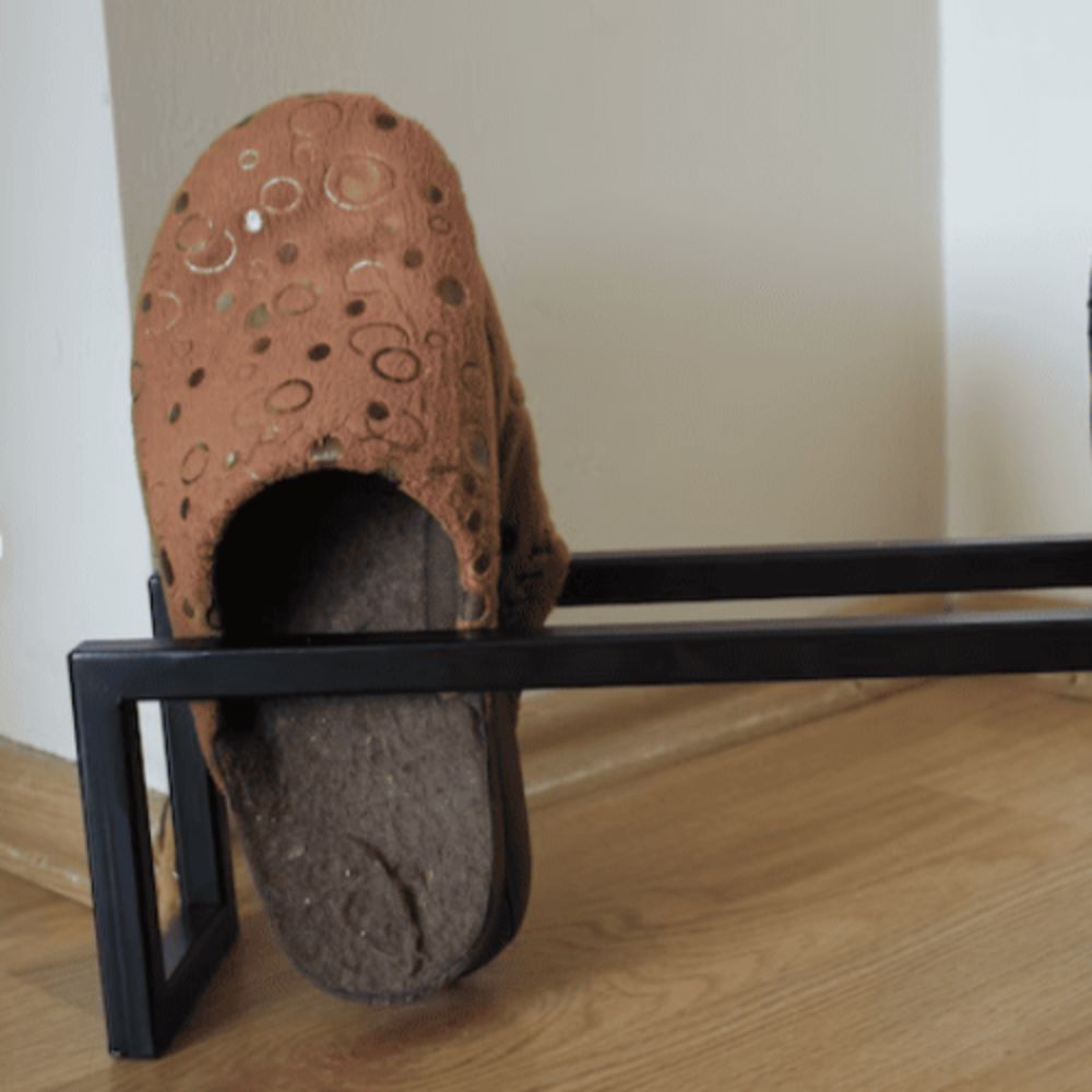 Ahşap zeminde çekilmiş, minimalist siyah metal bir rafta duran tek bir çift kahverengi terlik.