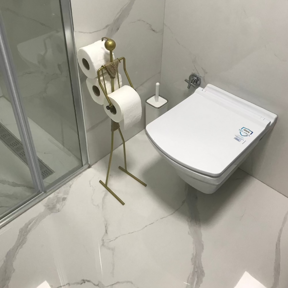 Şık, duvara monte bir tuvaletin yanında, birden fazla rulo tuvalet kağıdını tutan altın rengi şık, minimalist bir tuvalet kağıdı tutucunun bulunduğu modern bir banyo. Banyo lüks mermer bir yüzeye ve camla çevrili bir duşa sahiptir.