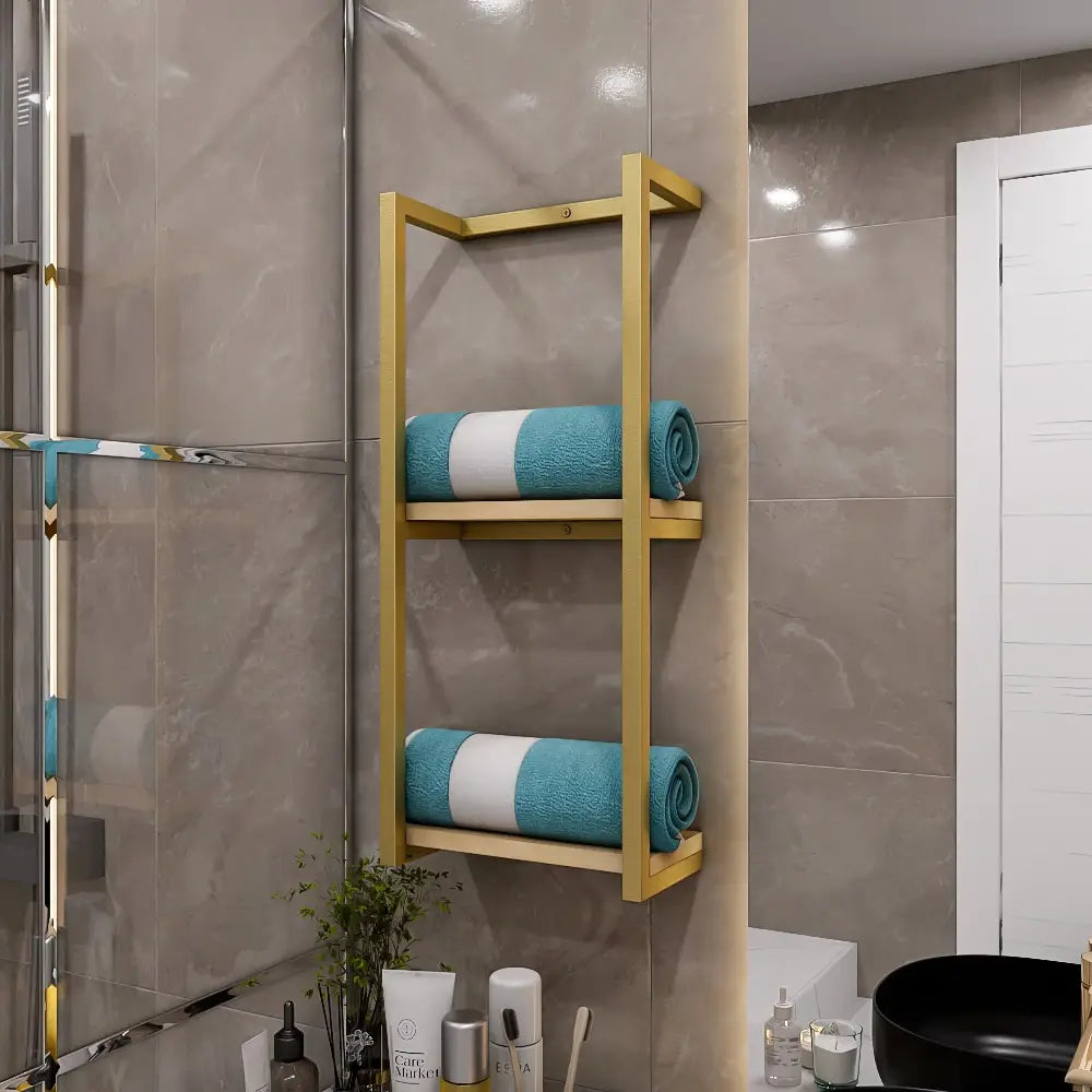 Koyu taş fayanslı bir duvara monte edilmiş, düzenli bir şekilde katlanmış mavi havluları tutan, modern bir estetik yaratan, açık ahşap tabanlı altın renkte metal banyo rafı.