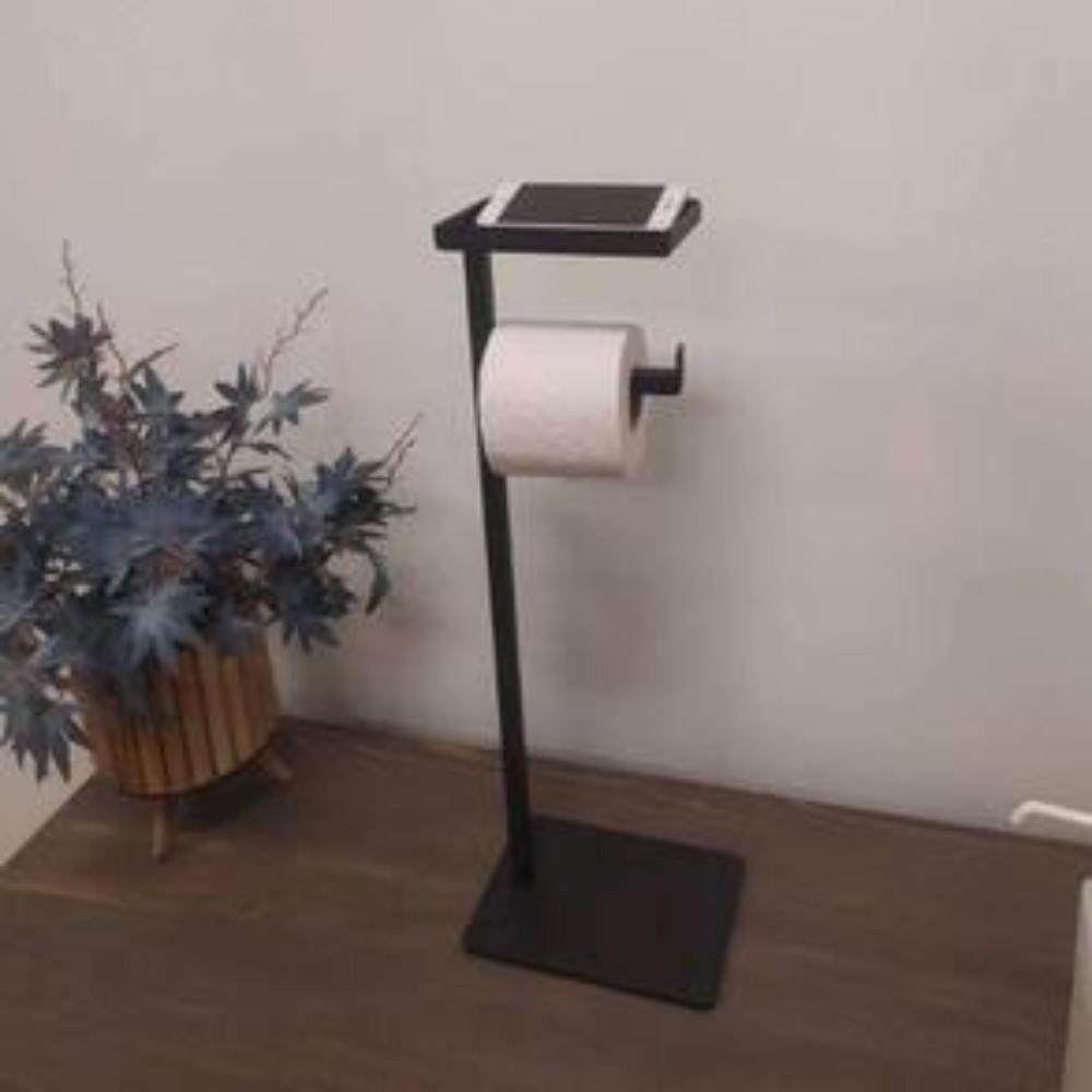 Ahşap bir yüzey üzerinde dekoratif bir bitkinin yanında duran, akıllı telefon tutan kompakt bir rafa sahip siyah, yere monte bir tuvalet kağıdı tutucusu.