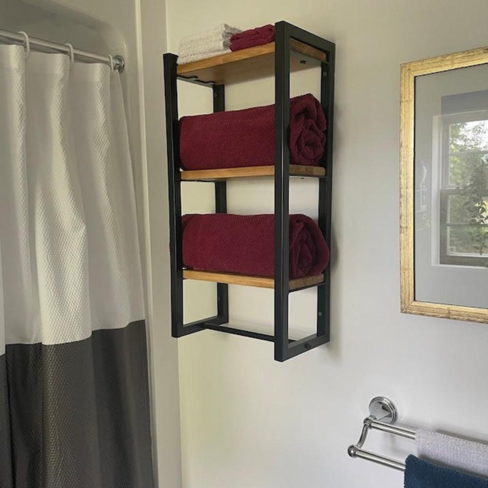 Modern bir banyo ortamında üç raflı banyo havluluğu. Düzenleyici, siyah metal çerçevelere ve ahşap raflara sahip olup, rulo bordo havluları düzenli bir şekilde tutmaktadır.