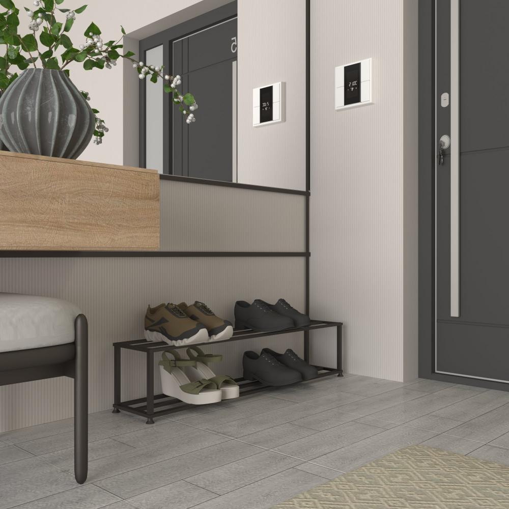 Şık, modern bir giriş alanına yerleştirilmiş minimalist siyah metal bir ayakkabılık. Rafın iki katında siyah klasik ayakkabılar ve gündelik spor ayakkabılar dahil çeşitli ayakkabılar bulunmaktadır. Şık alanda, bir vazoda yeşil bitki bulunan ahşap bir bank, dijital kilitli koyu renkli bir kapı ve çağdaş tasarımı yansıtan bir ayna bulunmaktadır. Açık renkli duvarlar ve zemin, ayakkabılığın işlevsel zarafetini vurgulayan temiz ve davetkar bir atmosfer yaratmaktadır.