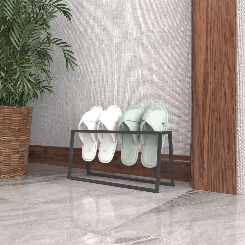 Minimalist tasarıma sahip siyah metal bir terliklik, içinde beyaz ve mint yeşili terlikler bulunduruyor. Ahşap bir duvarın ve saksı bitkisinin yanında, parlak mermer bir zeminin üzerinde duruyor.