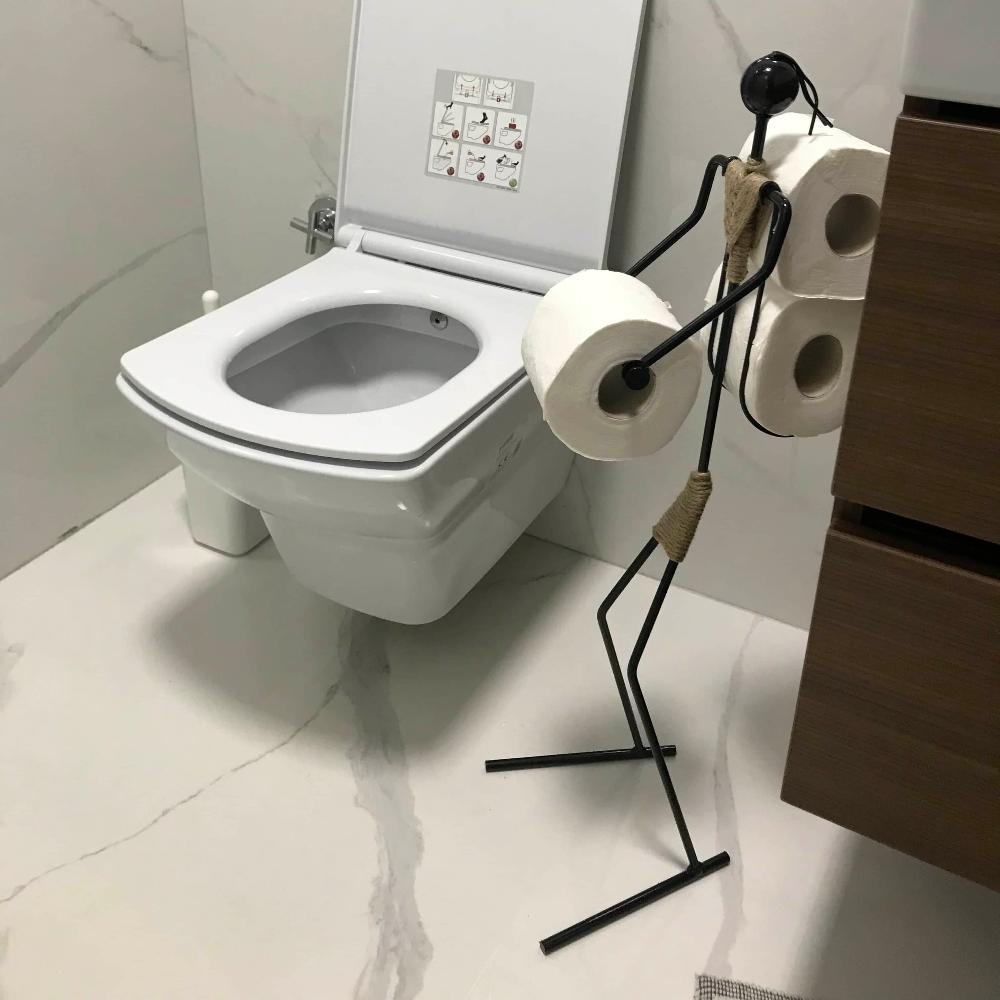 Mermer zemin ve duvar fayanslarına sahip bir banyoda modern beyaz bir tuvaletin yanına yerleştirilmiş, üç rulo tuvalet kağıdını tutan, figür şeklinde minimalist siyah metal tuvalet kağıdı tutucu.