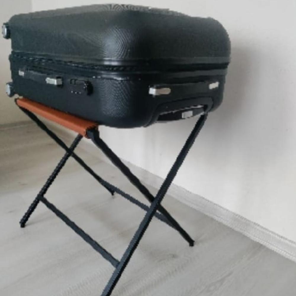 Siyah sert kabuklu bir bavul, deri üst yüzeyli katlanabilir metal bir bavul rafının üzerinde güvenli bir şekilde duruyor ve nötr bir iç mekanda konumlandırılmıştır.