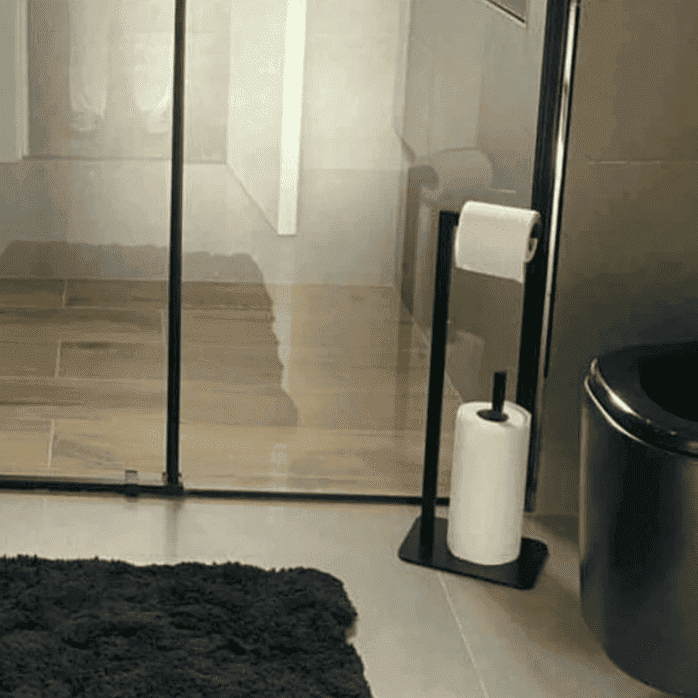 Cam bir duşakabin yanında, pelüş koyu bir kilim ve zarif iç mekanlarla tamamlanan, siyah bir tuvalet kağıdı tutucu standının bulunduğu bir banyo düzeni.