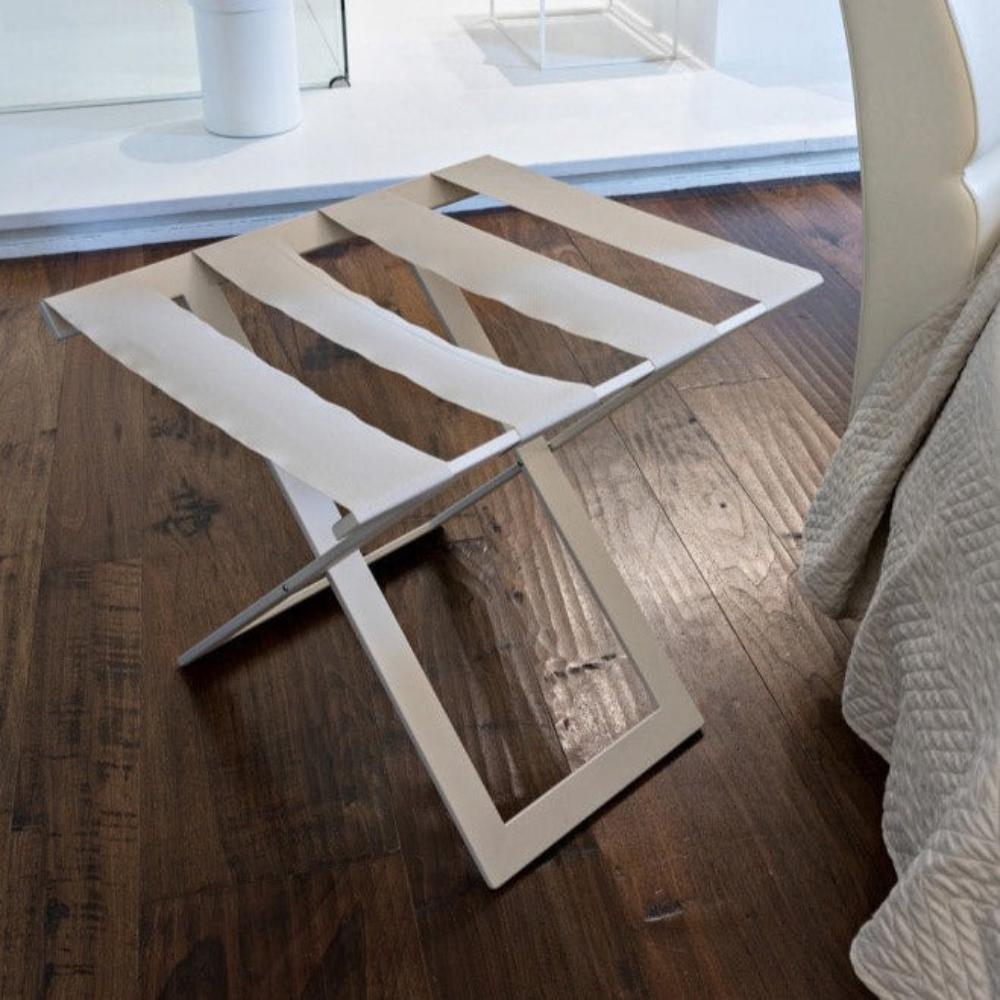 Minimalist bir yatak odası düzeninde, koyu ahşap zemine, krem rengi bir sandalyenin yanına yerleştirilmiş, sağlam kumaş kayışlara sahip modern bej metal bir bagaj rafı.