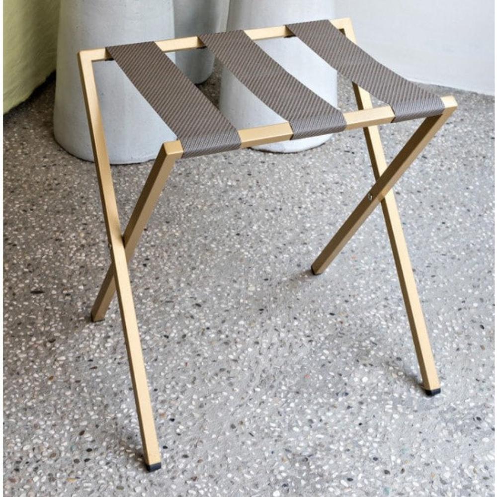 Terrazzo bir zemine yerleştirilmiş, sağlam ve minimalist bir tasarım sergileyen, kahverengi kumaş kayışlı altın rengi katlanabilir metal bagaj rafı.