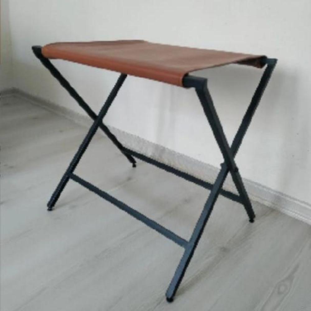 Hakiki deri üst kısmı olan katlanabilir metal bir bavul sehpası, ahşap bir zeminde minimalist bir iç mekanda sergileniyor.