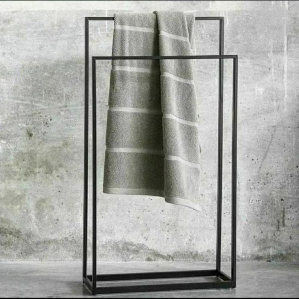 Modern, dokulu gri bir fon önünde sergilenen, üst barından düzenli katlanmış bir havlunun sarktığı, çağdaş banyo dekoru için minimalist tasarım ve işlevselliği sergileyen ayaklı siyah metal havluluk.