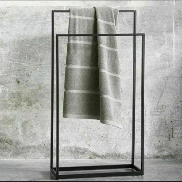 Modern, dokulu gri bir fon önünde sergilenen, üst barından düzenli katlanmış bir havlunun sarktığı, çağdaş banyo dekoru için minimalist tasarım ve işlevselliği sergileyen ayaklı siyah metal havluluk.