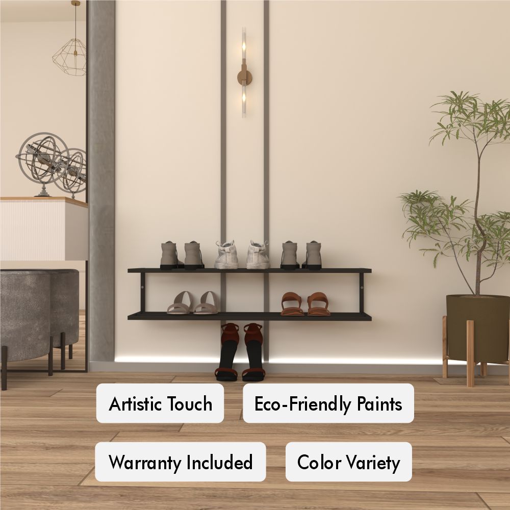 Wall-mounted black metal two-tier shoe storage shelf holding multiple pairs of shoes, with boots on the floor below. The background shows a minimalist interior with beige walls, a tall green plant on the right, and a modern industrial-style pendant light. Decorative labels below the shelf read "Artistic Touch," "Eco-Friendly Paints," "Warranty Included," and "Color Variety."