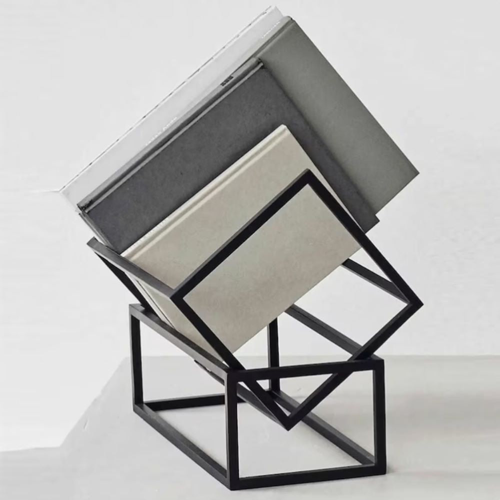 Modern, minimalist tasarıma sahip, üzerinde bir kitap yığını bulunan siyah metal bir kitap rafı, beyaz bir yüzeyin üzerine yerleştirilmiş.