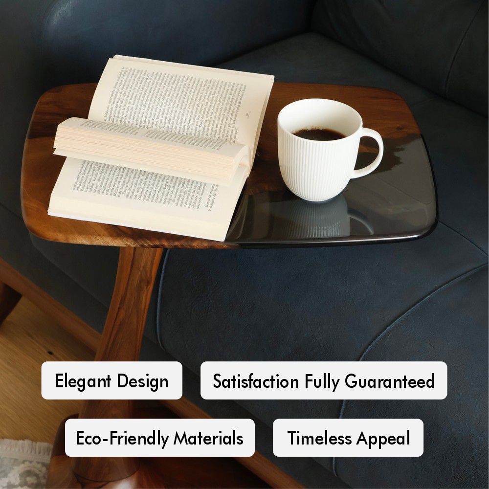 A close-up of a walnut wood side table with a glossy black resin top, featuring a coffee cup and an open book on a navy sofa.